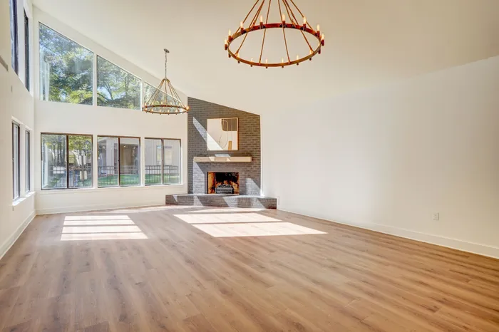 Spacious sunlit living room featuring large windows, sleek wood flooring, and a cozy brick fireplace.