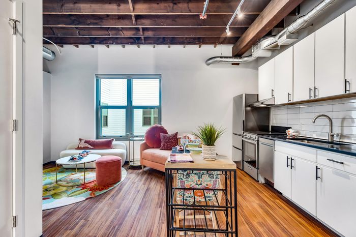 A contemporary living space featuring exposed wooden beams on the ceiling. The area is adorned with a white couch, pink ottomans, and a vibrant floral rug. Adjacent to the seating area is a modern kitchen with white cabinetry, stainless steel appliances, and a subway tile backsplash.