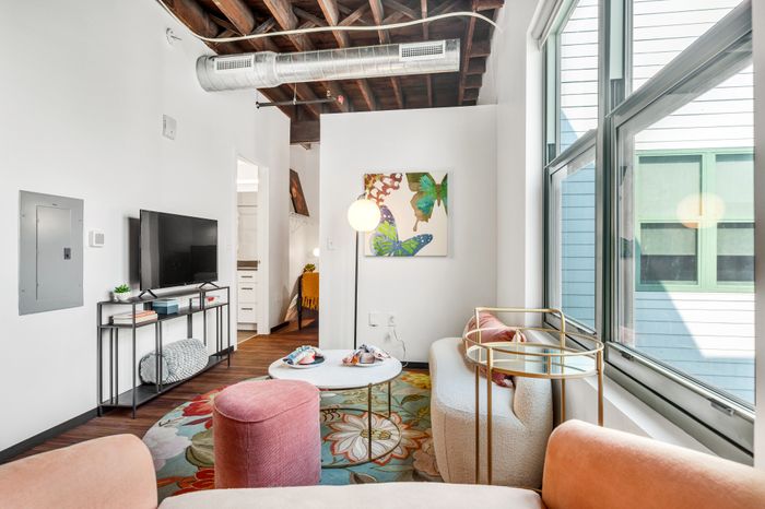 Modern living area with wooden ceiling beams and an exposed duct. A wall-mounted TV is placed beside a metallic artwork, and a white coffee table sits on a colorful rug. Nearby is a corridor leading to another room, illuminated by a pendant light.