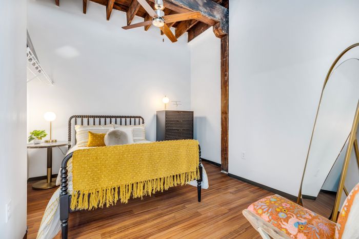 A cozy bedroom featuring wooden ceiling beams, a patterned white bed with yellow fringed blanket and complementary pillows. A standing arch lamp complements the space, and a desk with a white lamp is positioned against the wall.