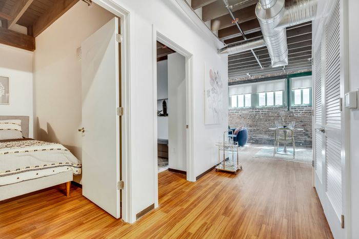 View of the entry hallway leading to the kitchen and living space, with doors on either side showcasing the two bedrooms. Hardwood flooring extends throughout, and the walls are adorned with art pieces. A glimpse into the living area reveals a blue armchair and glass table