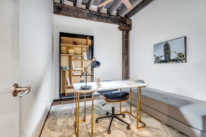 Contemporary workspace with exposed wooden beams. A white desk stands adjacent to a wooden bookshelf filled with various items. A modern chair is positioned by the desk, and a framed image of an airplane hangs on the wall.