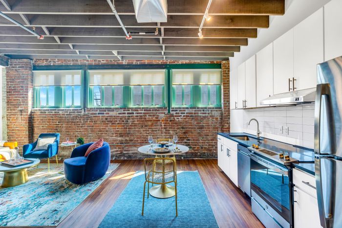 An open-concept living space highlighted by rustic wooden beams overhead. The kitchen showcases white cabinetry with modern fixtures, a stainless-steel oven, and a countertop stove. A white subway tile backsplash complements the dark countertops. The living area is anchored by a vibrant area rug and features plush blue seating arrangements, a round dining table with chic chairs, and large windows with brick detailing that allow ample natural light.