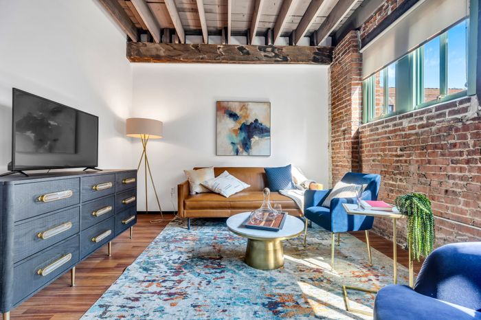 An open-concept living space highlighted by rustic wooden beams overhead. The kitchen showcases white cabinetry with modern fixtures, a stainless-steel oven, and a countertop stove. A white subway tile backsplash complements the dark countertops. The living area is anchored by a vibrant area rug and features plush blue seating arrangements, a round dining table with chic chairs, and large windows with brick detailing that allow ample natural light.