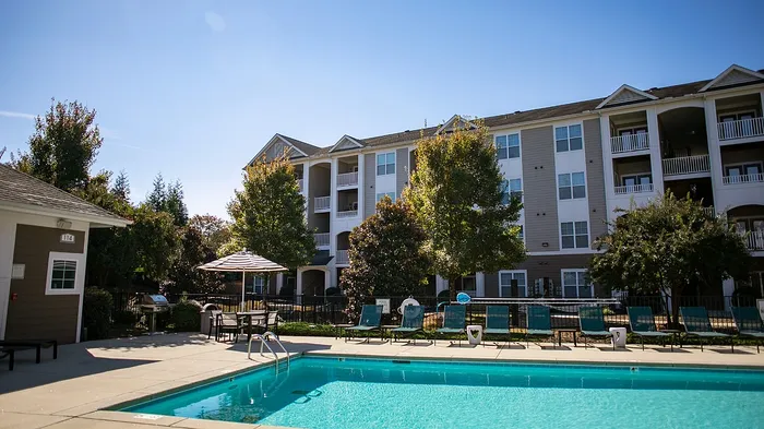 Sunny apartment complex with a sparkling outdoor pool, surrounded by lush trees and stylish loungers, perfect for relaxation and leisure.