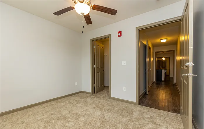 Inviting apartment room featuring plush carpeting and a modern ceiling fan, with an inviting view leading to a warmly lit hallway.