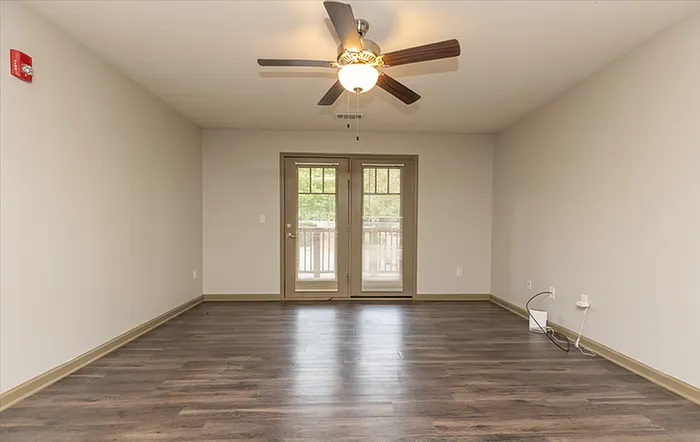 Spacious living room with elegant hardwood flooring and ceiling fan, featuring large double doors leading to a serene balcony view.