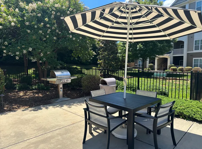 Charming patio setup with a stylish striped umbrella, modern table, and chairs, perfect for outdoor dining. Features a sleek stainless steel grill, surrounded by lush greenery and a luxurious apartment complex backdrop.