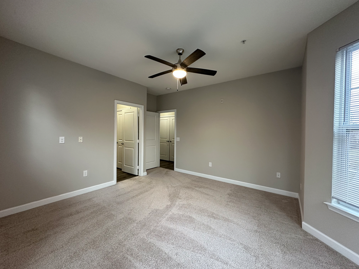 Bright and spacious bedroom featuring neutral walls, plush carpeting, and modern ceiling fan next to large windows for natural light.
