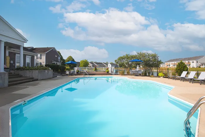 Inviting outdoor pool area with relaxing lounge chairs and a scenic backdrop of charming residential buildings and vibrant greenery under a bright blue sky.