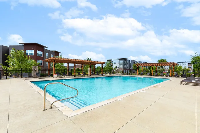 Inviting outdoor swimming pool with sleek loungers and modern pergolas, set against stylish apartment buildings under a bright blue sky.