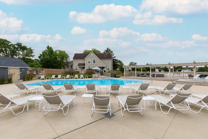 Inviting outdoor pool area with modern lounge chairs, surrounded by lush greenery and charming residential homes under a bright blue sky.