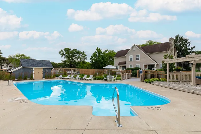 Inviting outdoor pool area with comfortable lounge chairs, surrounded by lush greenery and a charming residential house under a vibrant blue sky.