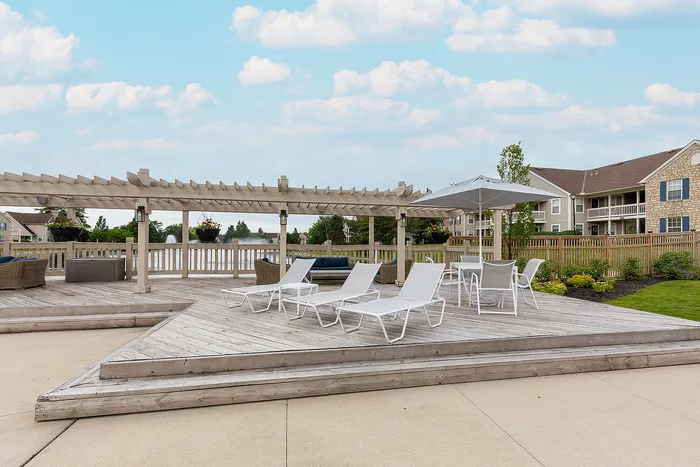 Charming outdoor deck with lounge chairs, pergola, and dining area set against a serene backdrop of landscaped gardens and modern apartments.