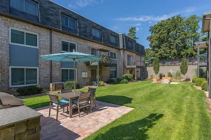 Inviting apartment courtyard with lush green lawn, patio furniture, and umbrella under a clear blue sky.