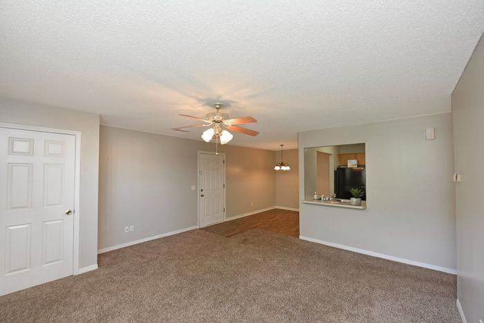 Bright open-concept living area with beige carpeting, ceiling fan, and view into the kitchen.