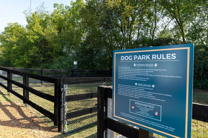 A welcoming entrance to a dog park with a sign displaying dog park rules, surrounded by lush greenery.
