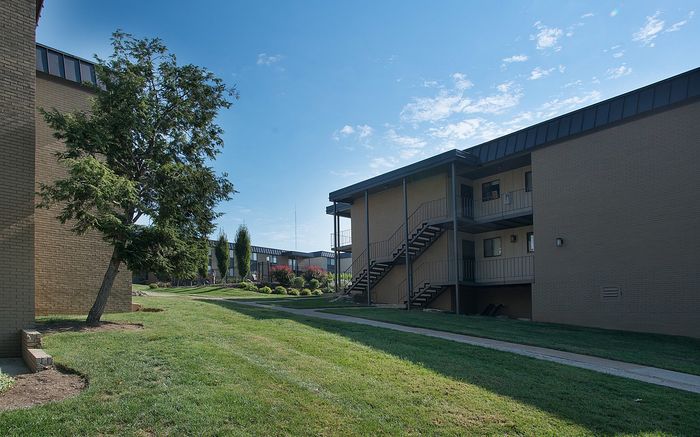 Exterior view of a brick apartment building with staircases and lush green landscaping under a bright blue sky.