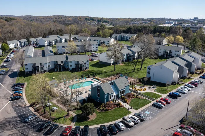 Charming aerial view of a contemporary apartment complex with lush green lawns, a refreshing swimming pool, and ample parking, nestled in a tranquil suburban setting.