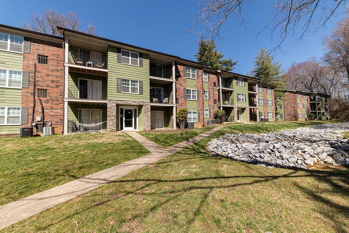 Charming apartment complex with brick and green exterior, featuring balconies and well-maintained grassy pathways under a clear blue sky.