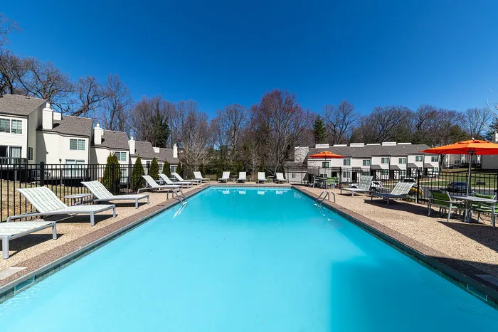 Inviting outdoor pool surrounded by sun loungers and vibrant orange umbrellas, set against a backdrop of charming townhouses and clear blue skies.