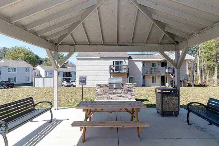 Charming outdoor common area with a covered seating pavilion, picnic table, and grill, offering serene views of the apartment community.