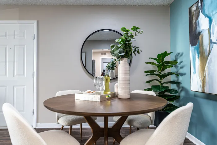 Elegant dining area with a round wooden table set with wine and glasses, complemented by plush chairs, a vibrant green plant, and a modern art piece, creating a chic and welcoming ambiance.