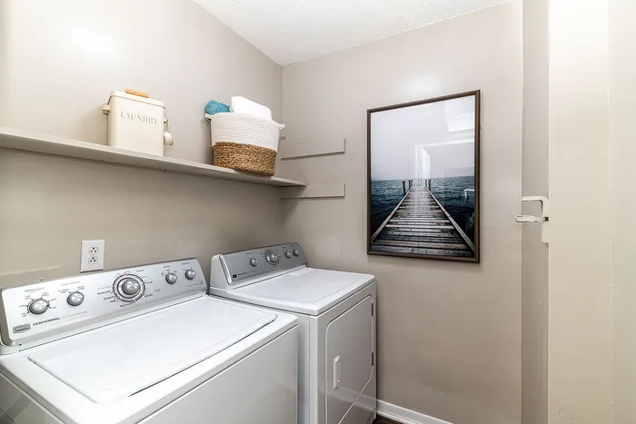 Charming laundry room featuring modern washer and dryer, elegant shelving with decor, and serene coastal artwork.