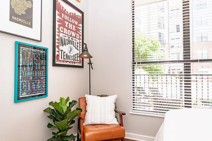 Bright and inviting apartment corner with stylish wall art, cozy leather chair, and lush indoor plant, flooded with natural light through large windows.