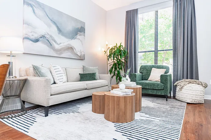 Bright and modern living room with elegant decor, featuring a plush green armchair, cozy beige sofa, and stylish abstract artwork. The room is complemented by natural light streaming through large windows and vibrant indoor plants.