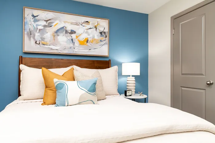 Modern bedroom with a calming blue accent wall, featuring a cozy bed adorned with colorful pillows, elegant abstract artwork, and a stylish table lamp.