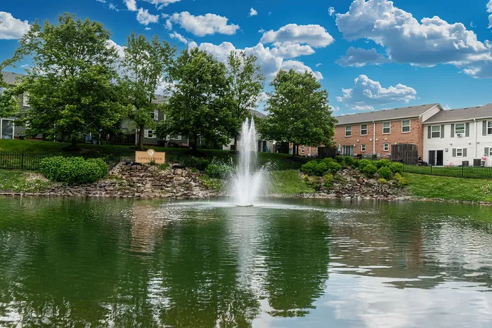 Charming apartment complex with picturesque pond view, fountain, and lush green surroundings under a bright blue sky.