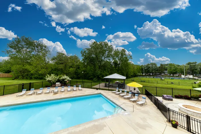Luxurious swimming pool area with sun loungers, shaded seating, and lush green surroundings under a bright blue sky with fluffy clouds.