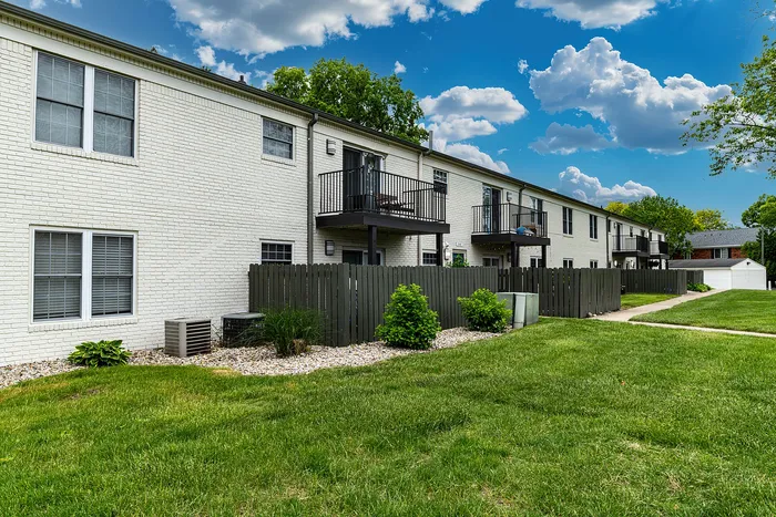 Charming white-brick apartment complex with private balconies, surrounded by lush green lawns and a clear blue sky.