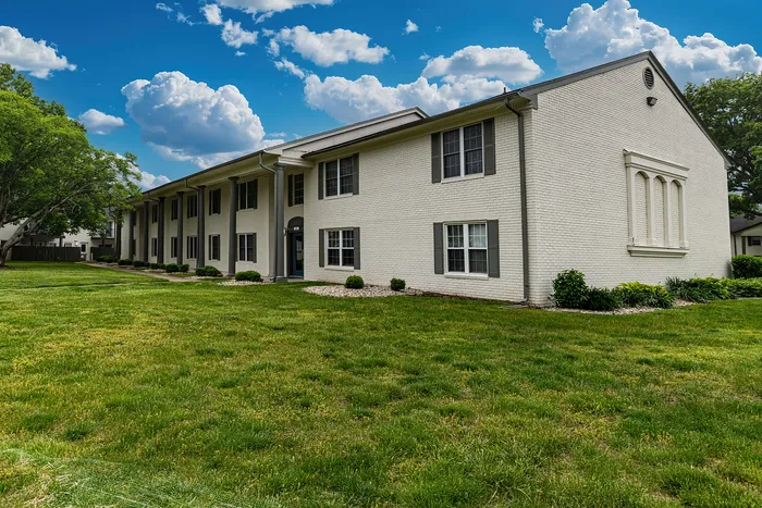 Charming white brick apartment building with manicured lawns, surrounded by lush trees and a vibrant blue sky.