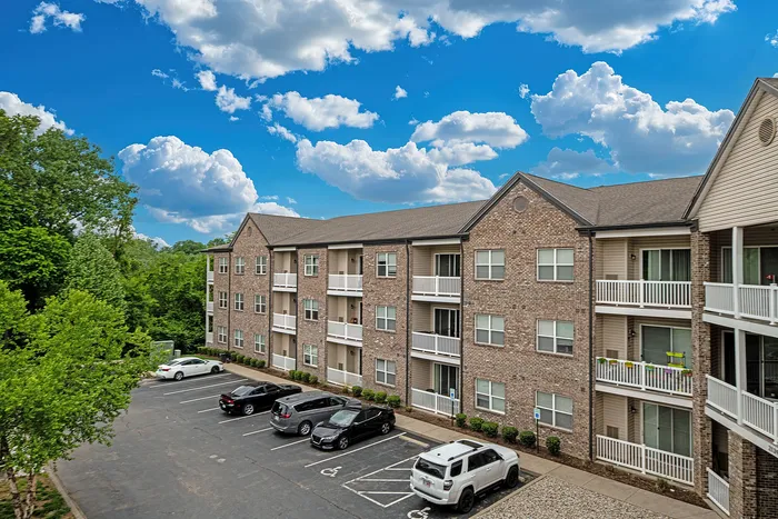 Charming three-story apartment building with brick fa&ccedil;ade, featuring spacious balconies and surrounded by lush greenery under a vibrant blue sky.