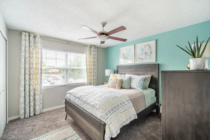 Bright and inviting bedroom with modern decor, featuring a plush bed, stylish ceiling fan, and large window with elegant curtains.