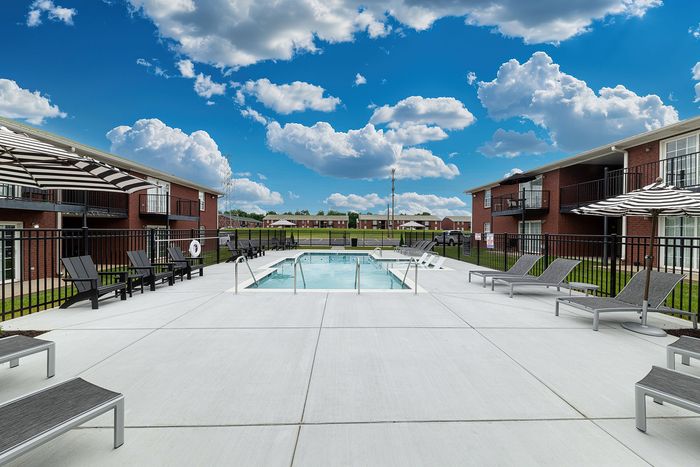 Inviting outdoor pool area with modern sun loungers, shade umbrellas, and vibrant blue skies, surrounded by charming brick apartment buildings.
