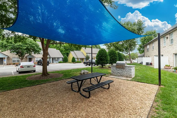 Charming outdoor area with a shaded picnic table and a modern grill, surrounded by lush greenery and picturesque townhouses.