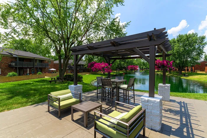 Charming outdoor patio with elegant pergola overlooking a serene pond, featuring stylish seating and vibrant hanging flowers.