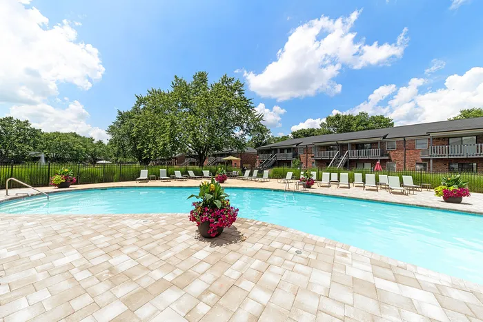 Inviting apartment pool area with sun loungers, vibrant flowers, and lush greenery under a bright blue sky.