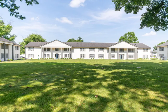 Charming colonial-style apartment building surrounded by lush green lawn, featuring elegant pillars and bright windows, perfect for serene living.