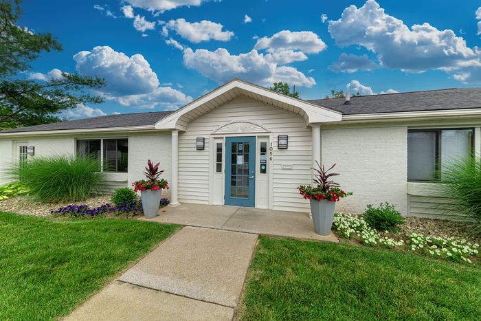 Charming single-story home entrance with elegant landscaping, vibrant flower pots, and a striking blue door against a picturesque sky.