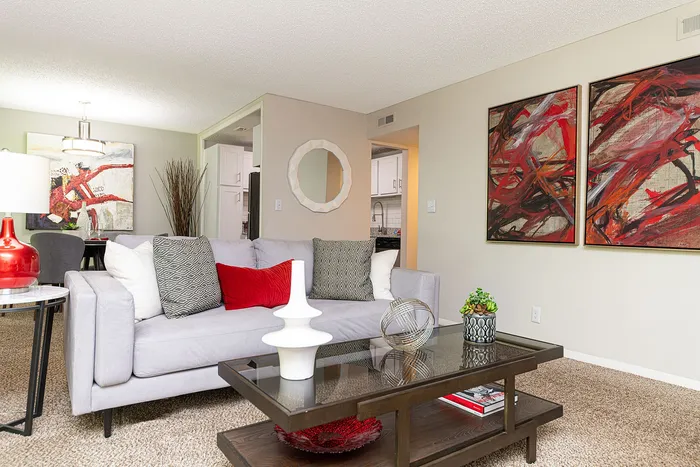 Elegant living room with modern gray sofa, vibrant red and white decor, abstract art, and open view to a cozy kitchen.
