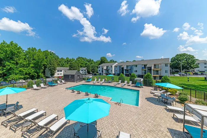 Inviting apartment community with a sparkling outdoor pool, relaxing sun loungers, and vibrant blue umbrellas under a clear sky.