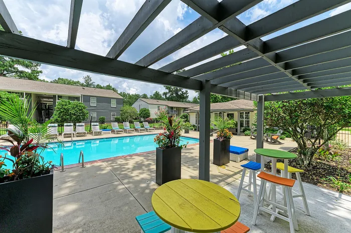 Vibrant outdoor swimming pool area with colorful seating under a pergola, surrounded by lush greenery and modern apartment buildings.