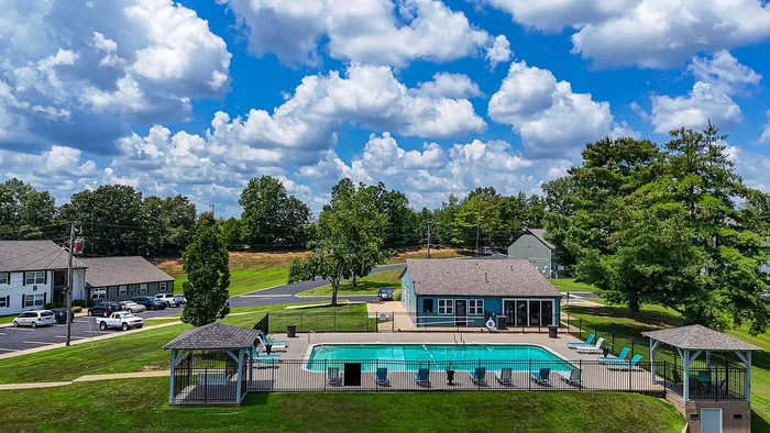 Inviting apartment complex with a refreshing swimming pool, surrounded by lush green landscape under a vibrant blue sky with fluffy clouds.