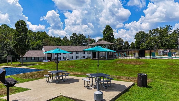 Charming outdoor space with vibrant green lawn, picnic tables under turquoise umbrellas, and a fenced pool area, ideal for relaxation and leisure.
