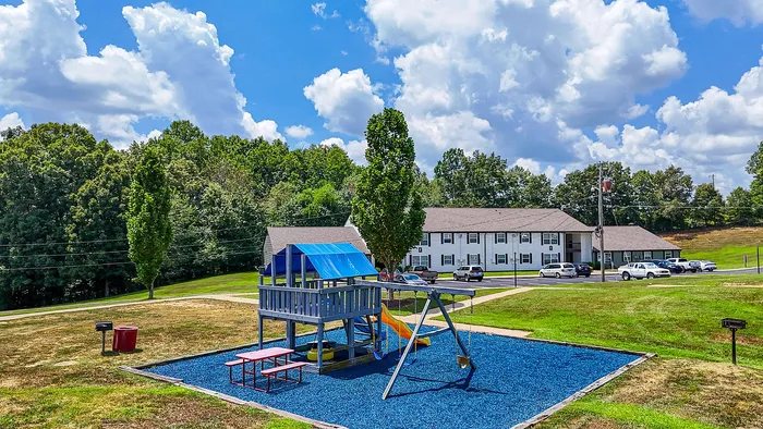 Charming apartment complex with a vibrant playground, surrounded by lush green trees and open skies.