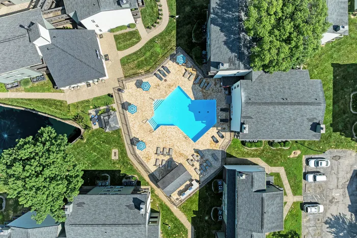 Aerial view of a luxurious apartment complex featuring a large, sparkling blue swimming pool surrounded by stylish lounge chairs and vibrant umbrellas, nestled among neatly landscaped green spaces and modern buildings.